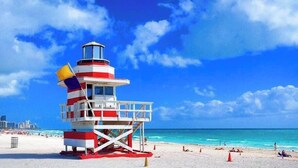 Un phare à plusieurs niveaux, aux couleurs rouge et blanc, situé sur une plage bordée d’un ciel bleu clair et d’eaux turquoise.