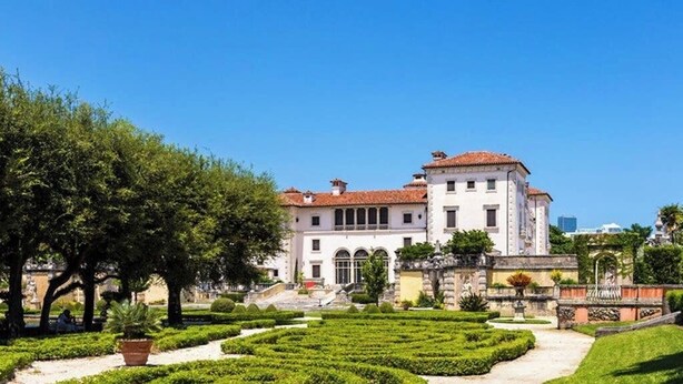 Un grand bâtiment blanc surmonté d’un toit recouvert de tuiles rouges, entouré d’un jardin bien entretenu avec un chemin central et des arbres.