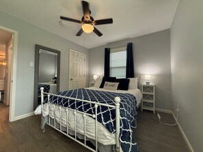 Une chambre à coucher comprenant un lit, des tables de chevet, un ventilateur de plafond, une fenêtre avec des rideaux et une porte.