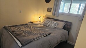 Ein Schlafzimmer mit einem Bett, einer Nachttischlampe, einem Fenster mit Jalousien und einem an der Wand hängenden Bild.