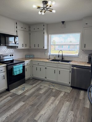 A modern kitchen with white cabinets, a stainless steel dishwasher, and a large window with blinds. - Serene 2-bedroom house with water view in charming Venice (Venice)