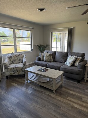 A living room with a grey sofa, armchair, and coffee table. There are two windows with curtains, a ceiling fan, and a plant. - Serene 2-bedroom house with water view in charming Venice (Venice)