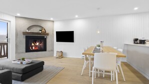 Una sala de estar moderna con chimenea, un comedor con mesa de madera y sillas blancas, y una cocina con encimera y electrodomésticos blancos.