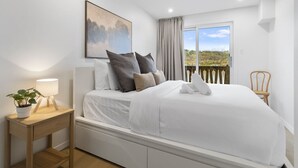 Una cama bien hecha con almohadas, una mesita de noche con una lámpara y una planta, una ventana con cortinas y vistas al exterior.