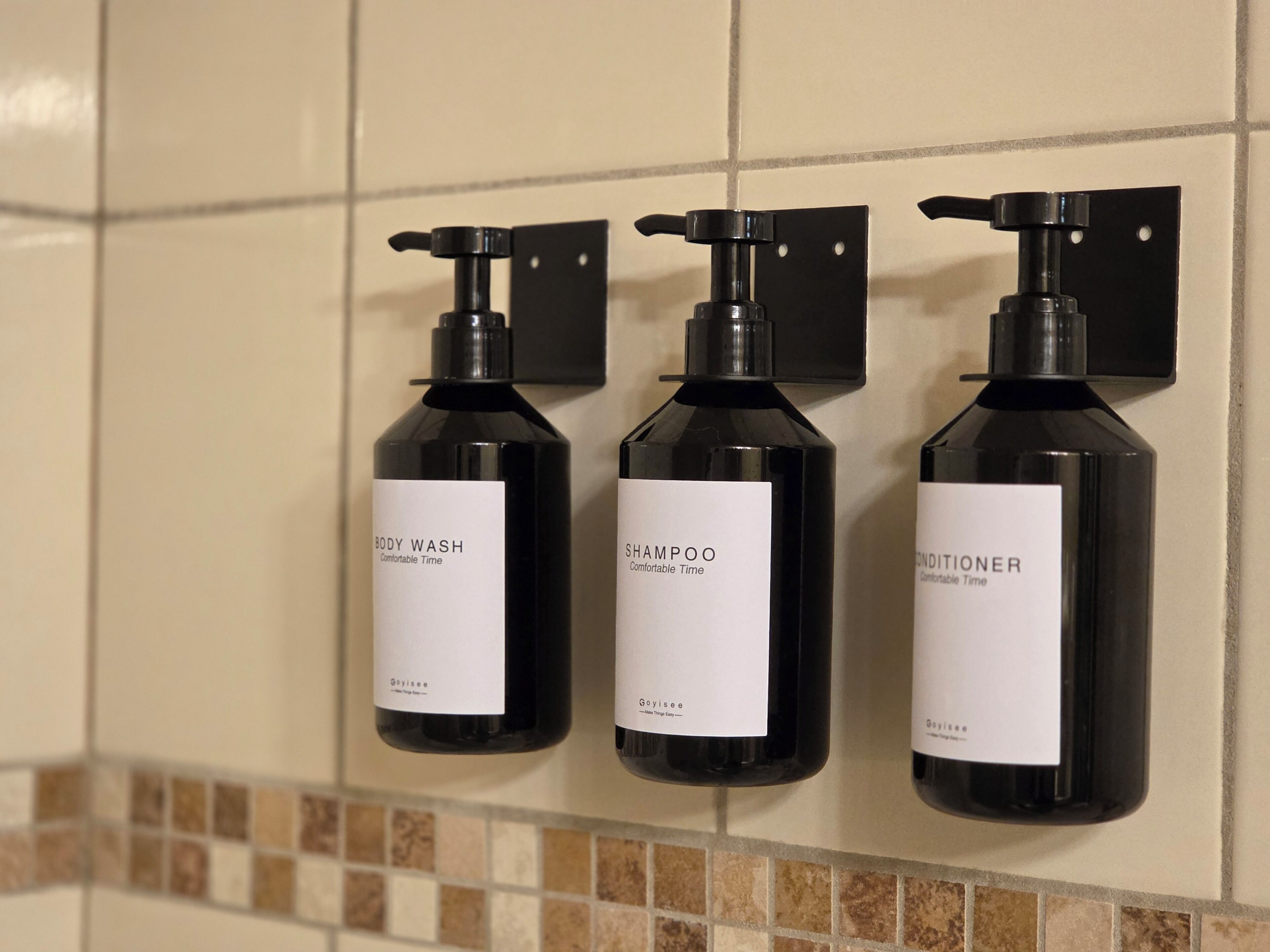 Three black bottles of body wash, shampoo, and conditioner mounted on a tiled wall.