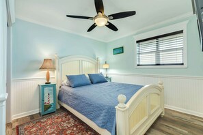 A bedroom with a bed, a ceiling fan, a window with blinds, a nightstand with a lamp, and a wall-mounted sign. - Direct Beach Access Surfside Oceanfront Home (Surfside Beach)