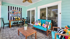 A covered patio with a teal-colored wall, a wooden table, a sofa with colorful cushions, and a ceiling fan. - House of Whimsy (Tybee Island)