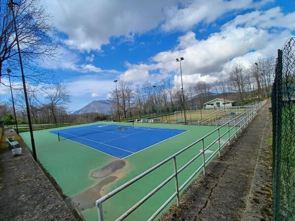 Una cancha de tenis al aire libre con superficie verde y azul, rodeada por una valla metálica y árboles.