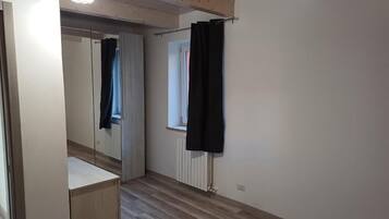 A bedroom with a wooden floor, a bed, a wardrobe, a mirror, and a window with curtains.