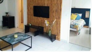 A modern living room with a flat-screen TV mounted on a wooden panel wall, a glass coffee table, and a bed with a floral bedspread.