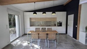 Une cuisine moderne avec des meubles blancs, une table à manger avec des chaises, des luminaires suspendus et une grande fenêtre.