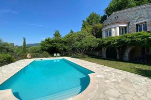 Ein rechteckiger Swimmingpool mit Steinplatten, umgeben von einer Steinterrasse, und ein Steinhaus mit Säulen und einem überdachten Bereich auf der rechten Seite.
