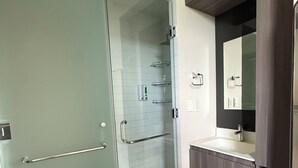 Un baño con mampara de vidrio, un lavamanos blanco, un mueble de tocador de madera oscura y un espejo sobre el lavamanos.