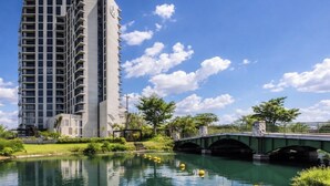 Un moderno edificio de gran altura con un cuerpo de agua reflectante al frente, un puente verde y abundante vegetación.
