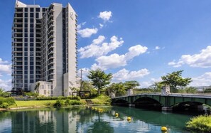 A modern high-rise building with a reflective water body in front, a green bridge, and lush greenery. - APARTMENT IN MERIDA YUCATAN (Mérida)