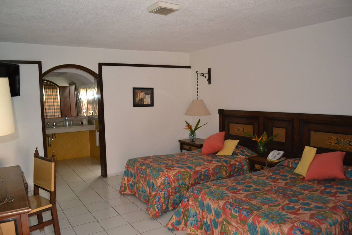 Habitación de hotel con dos camas, un escritorio, una silla, una mesita, una mesita de noche, una lámpara, un cuadro en la pared y un baño visible a través de un arco.