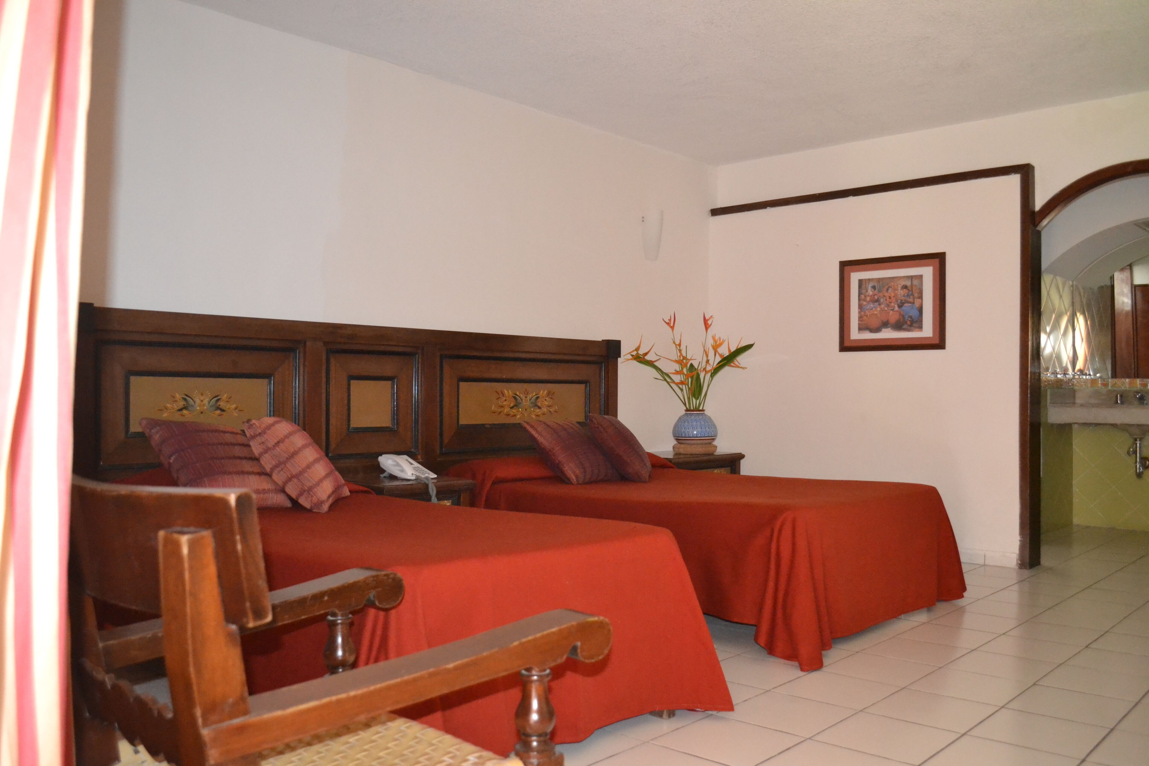A hotel room with two beds, a wooden headboard, a bedside table with a lamp, a vase with flowers, and a framed picture on the wall.