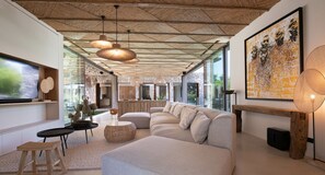 A modern living room with a large sectional sofa, a TV, and a dining area with a stone wall and wooden ceiling. - New 362 m² fully equipped villa, located in Ramatuelle (Ramatuelle)