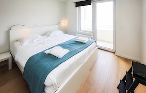 Ein ordentlich bezogenes Bett mit weißen Laken und einer türkisfarbenen Decke, ein Nachttisch, ein Fenster mit Blick und ein Schreibtisch mit Stuhl.