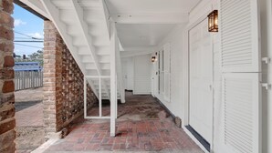 A covered porch with a brick wall, white siding, and a staircase leading to a door. - Fire & Oaks Estate (Tybee Island)