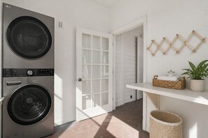 A laundry room with two stacked washers and dryers, a white door, a shelf with baskets, and a potted plant. - Fire & Oaks - The Oak Villa (Tybee Island)