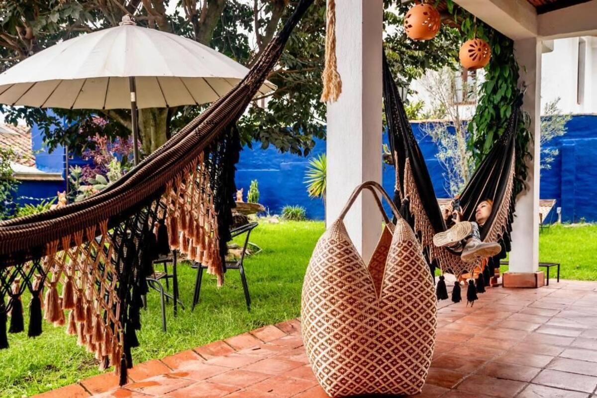 A hammock under a white umbrella, a wicker chair, and a patterned bag on a patio with a view of a blue wall and greenery.