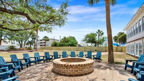 A circular brick fire pit surrounded by blue Adirondack chairs, with a brick patio and trees in the background. - Fire & Oaks - The Ember Villa (Tybee Island)