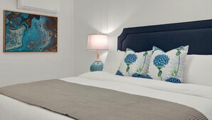A neatly made bed with a headboard, a bedside lamp, and a wall-mounted air conditioner. - Fire & Oaks - The Ember Villa (Tybee Island)