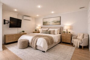 A bedroom with a bed, bedside tables, a TV, a painting, and a chair. - Fire & Oaks - The Ember Villa (Tybee Island)