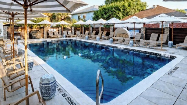 A swimming pool with lounge chairs and umbrellas, surrounded by a brick wall and greenery. - Fire & Oaks - The Ember Villa (Tybee Island)