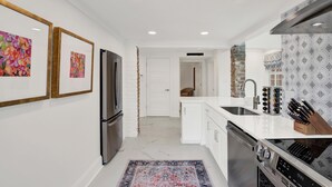 A modern kitchen with white cabinetry, a stainless steel refrigerator, a built-in oven, and a stone accent wall. - Fire & Oaks - The Ember Villa (Tybee Island)