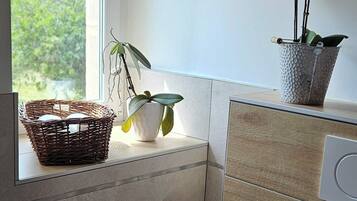 A bathroom with a window, a sink, a toilet, a trash bin, and a small potted plant.