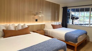 Habitación de hotel con dos camas, cabecero de madera, mesitas de noche, una ventana con cortinas e iluminación en el techo.