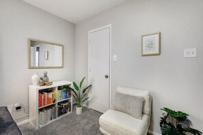A room with a white bookshelf, a white armchair, a door, and a framed picture on the wall. - Candleridge Casa - Newly Remodeled 4-bd Hidden Gem (Fort Worth)