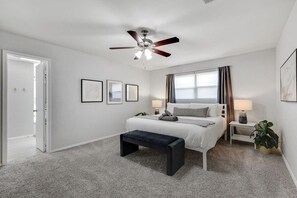 Un dormitorio con cama, mesitas de noche, ventilador de techo, ventana con cortinas e imágenes enmarcadas en la pared.