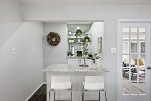 A kitchen with a granite countertop, white chairs, a hanging wreath, and a view through a glass door to a living area. - Candleridge Casa - Newly Remodeled 4-bd Hidden Gem (Fort Worth)