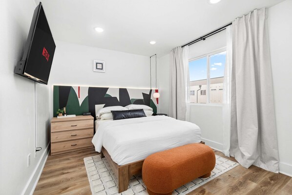 Ein modernes Schlafzimmer mit Bett, Wandfernseher, Holzkommode und einem Fenster mit Vorhängen.