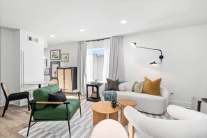Ein modernes Wohnzimmer mit einem grünen Sofa, einem weißen Sessel und einem grauen Sofa. Zwei runde Holztische, eine schwarze Stehlampe und eine weiße Kommode sind ebenfalls vorhanden.