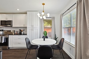 A modern kitchen with white cabinets, a stainless steel oven, and a dining area with a white table and black chairs. - Modern Comforts & Classic Charm w Pool, 5BD (Fort Worth)