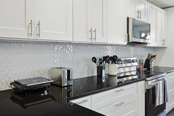 A modern kitchen with white cabinets, a black countertop, a toaster, a waffle maker, and a microwave. - Modern Comforts & Classic Charm w Pool, 5BD (Fort Worth)