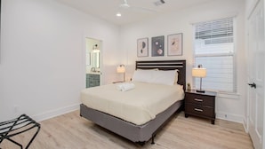Une chambre à coucher comprenant un lit, des tables de chevet, un ventilateur de plafond, une fenêtre avec des stores et un miroir.