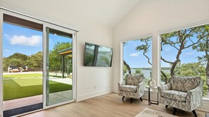 A modern living room with a large sliding glass door leading to a backyard, a flat-screen TV mounted on the wall, and two patterned armchairs. - Heavenly Retreat 5bd/5.5ba of Waterfront living (Gainesville)