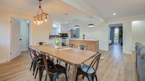 A spacious dining area with a long wooden table and six chairs, a hanging light fixture, and a kitchen in the background. - 4-bd Central Location w Pool, Patio, Firepit (Houston)