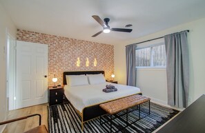 Ein Schlafzimmer mit Backsteinwand, Deckenventilator, Bett mit Kissen, einer Bank, einem Nachttisch und einem Fenster mit Vorhängen.