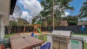 A backyard with a wooden table, chairs, a barbecue grill, a playset, and a wooden fence. - Spring Branch Paradise, 4-bd Backyard Pool Oasis (Houston)