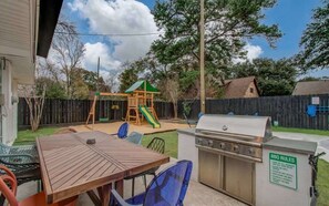 Ein Garten mit Holztisch, Stühlen, Grill, Spielplatz und Holzzaun.