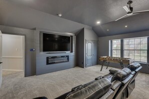 A home entertainment room with a flat-screen TV, a foosball table, and a reclining leather couch. - 5BR Oasis w/ Pool Steps From Lake Ray hubbard (Rowlett)