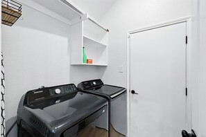 A laundry room with two Samsung washing machines, a white door, and a shelf with a bottle and a box. - 5-bd/2-ba Upscale Elegance Pool, Hot tub, bbq (Frisco)