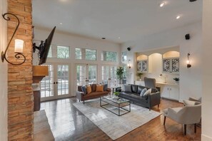 A spacious living room with a stone accent wall, a large window, a fireplace, and a modern fireplace mantel. - Dallas Gem 5 Bedrooms Home With Pool, Game Room (Dallas)