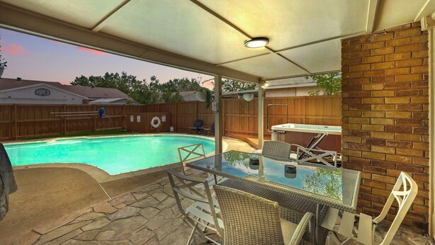 A covered patio with a glass table and chairs, a brick wall, and a backyard pool. - Modern & Chic 4-bd/2-ba Pool, Hot Tub, BBQ Grill (Carrollton)